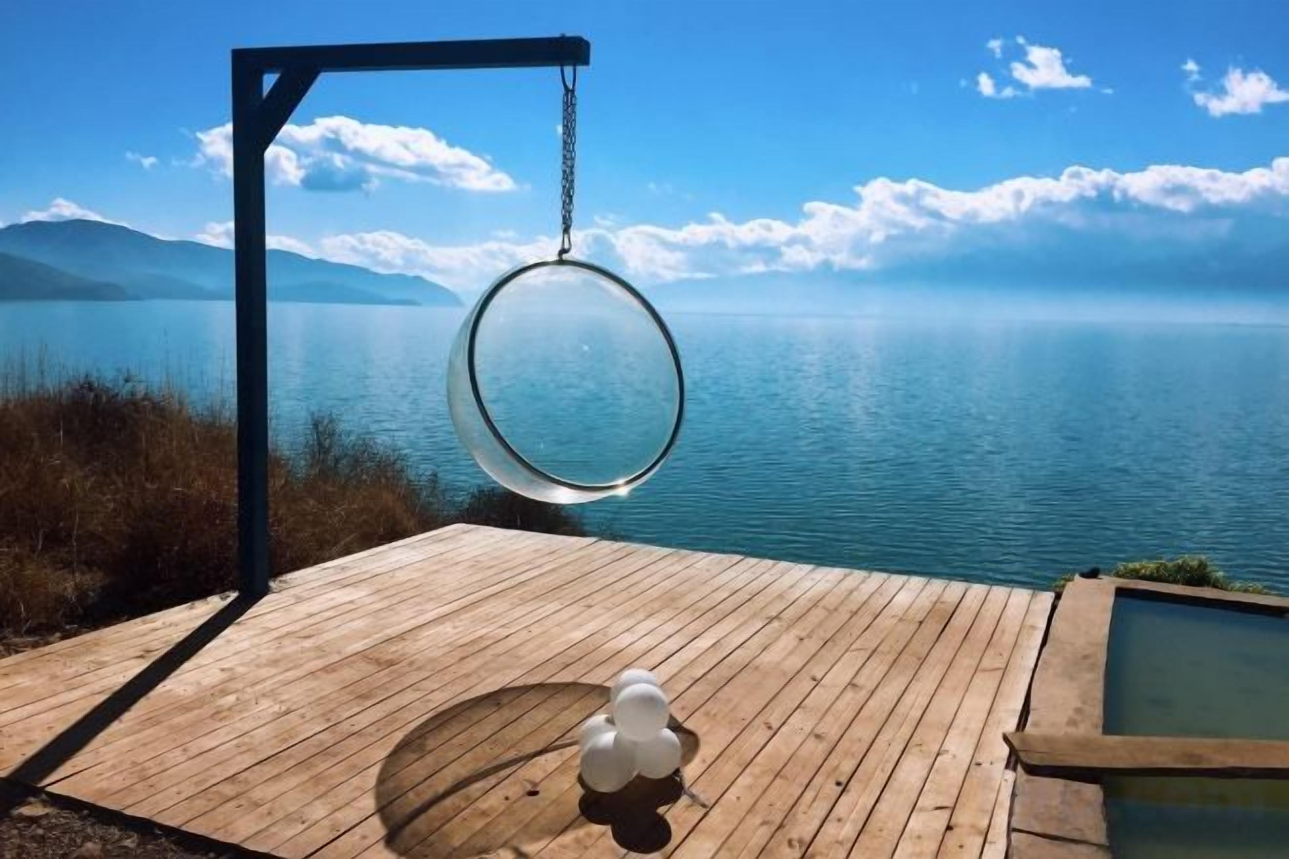 A wooden deck with a hanging swing overlooking a calm lake and mountains.