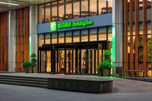 A Holiday Inn hotel entrance with a glass facade, a green sign, and a small outdoor seating area with planters. - Holiday Inn Beijing Airport Zone by IHG (Beijing)