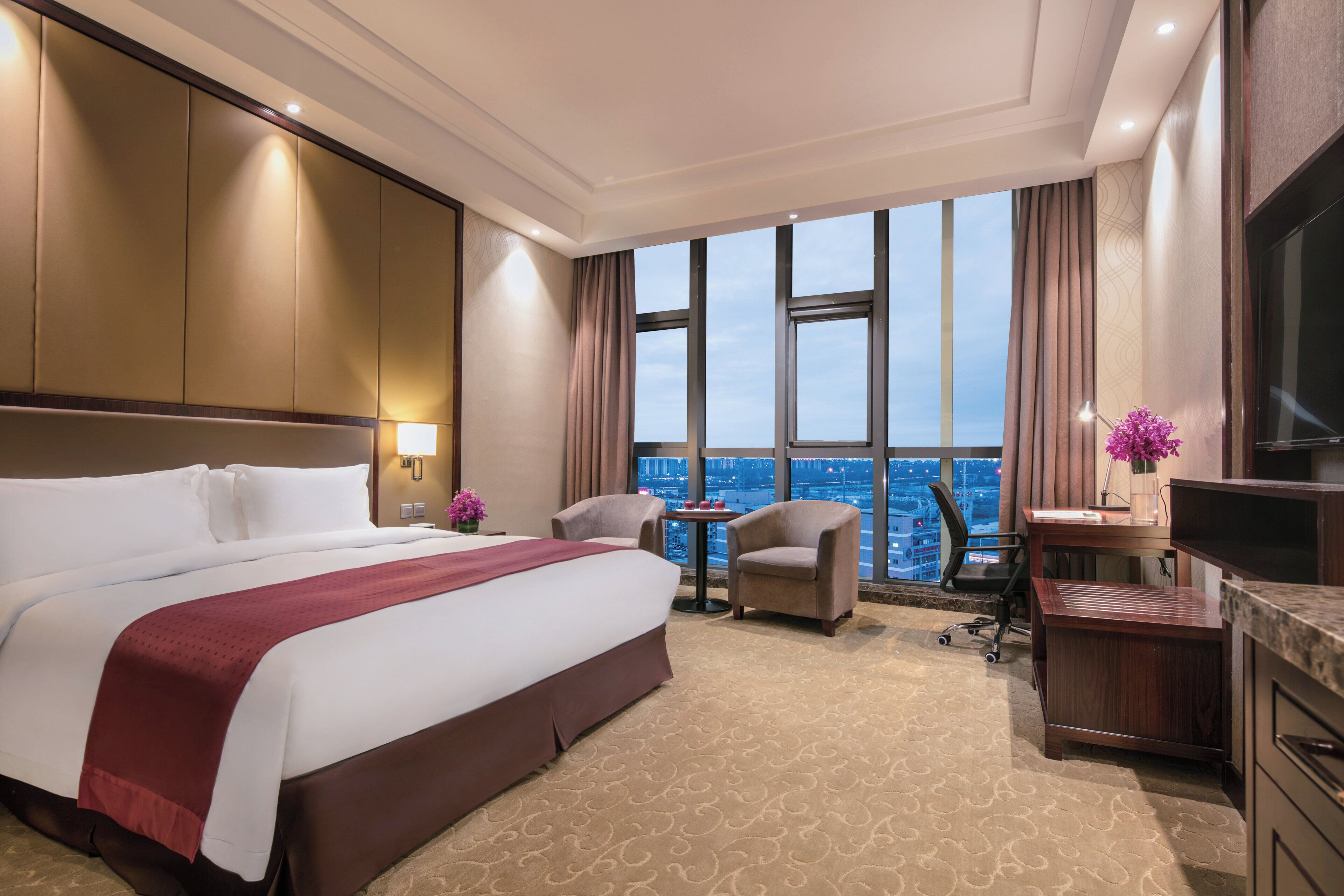 A hotel room with a large bed, two armchairs, a desk, a TV, and a view of the cityscape through large windows.