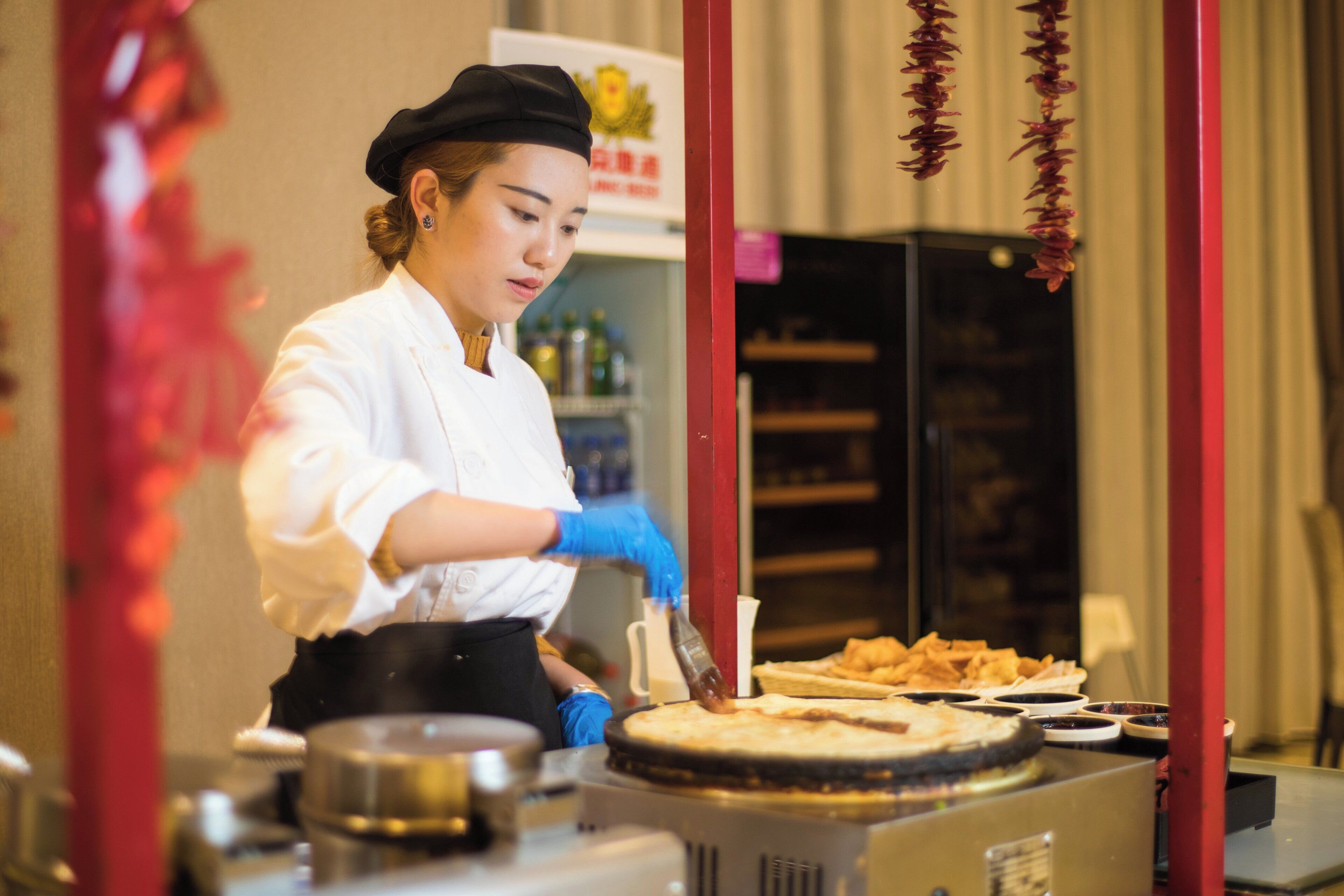 Une personne préparant des aliments sur un comptoir, avec une vitrine réfrigérée et du matériel de cuisine en arrière-plan.