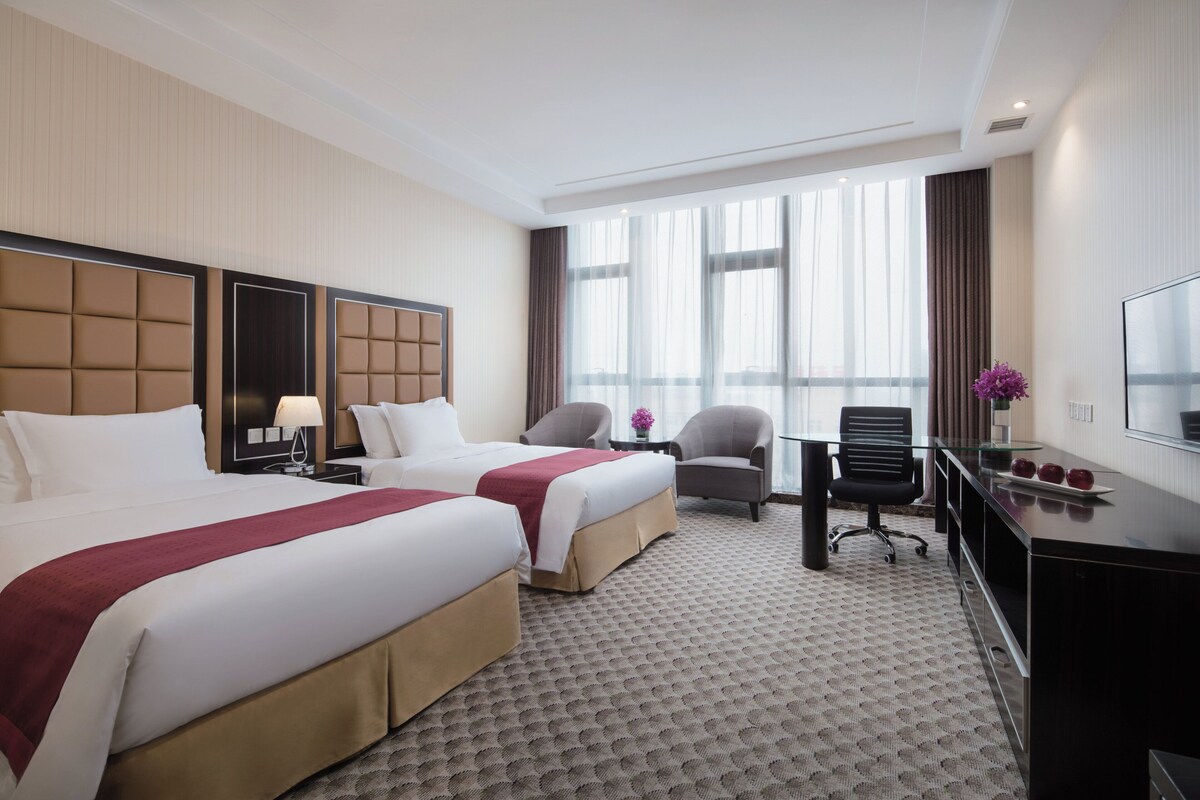 A hotel room with two beds, a desk, a chair, and a TV.