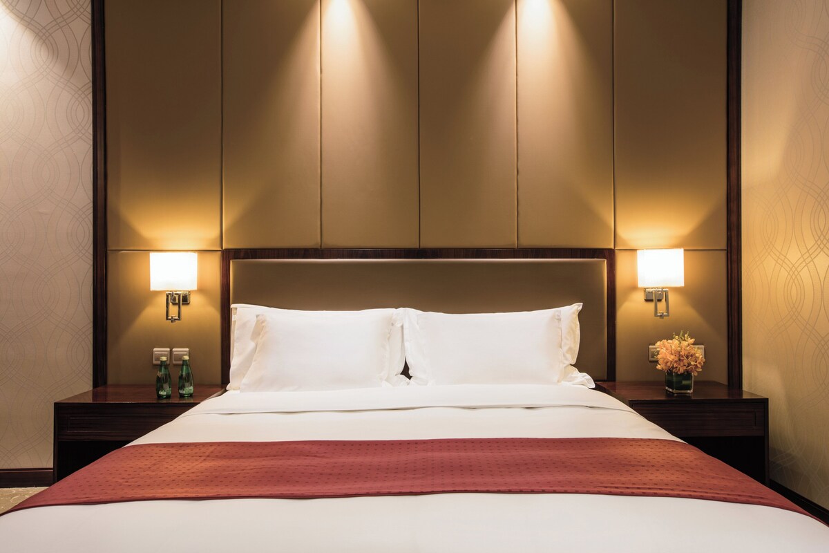 A hotel room with a large bed, two bedside lamps, a nightstand with a bottle, and a vase with flowers.