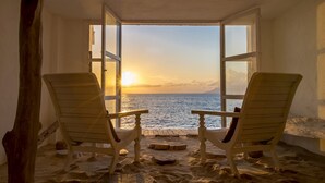 Dos sillas de madera mirando hacia una ventana con vista al mar al atardecer.