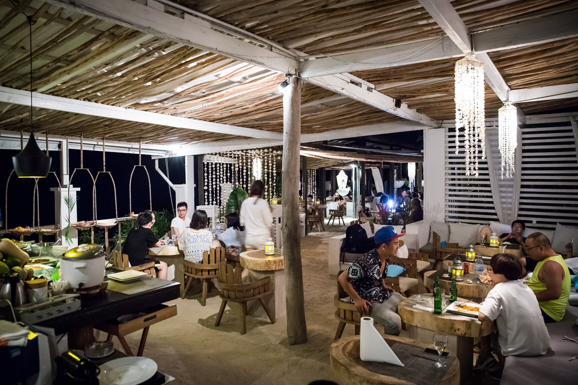 An outdoor dining area with wooden tables and chairs, surrounded by a rustic wooden structure with a thatched roof.