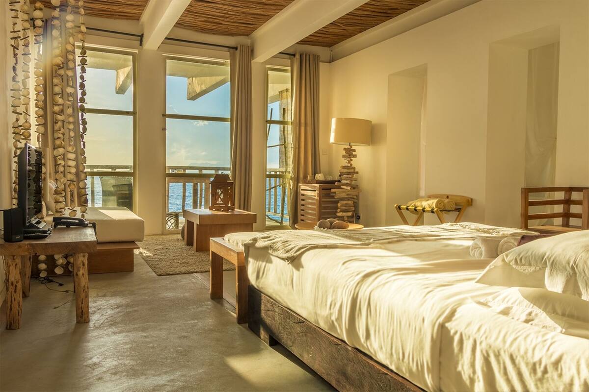 Un dormitorio con una cama grande, un escritorio con teléfono, una lámpara y vistas al mar a través de amplios ventanales.