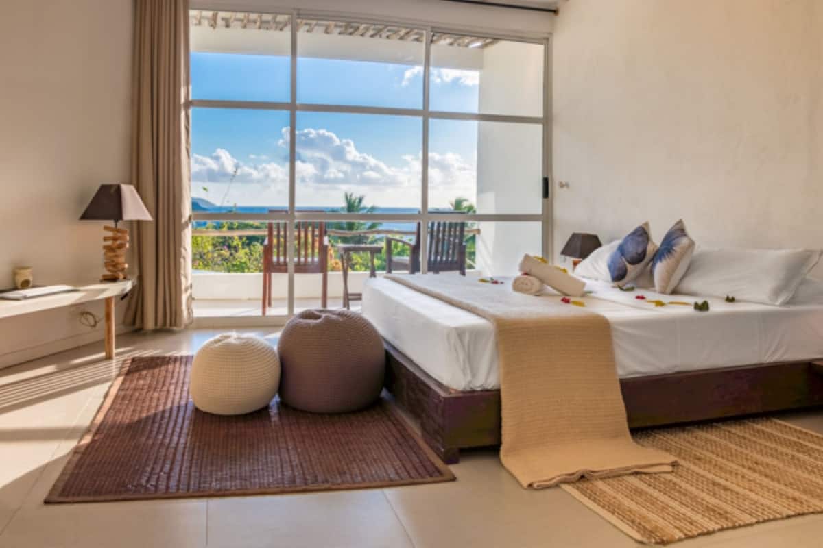 Un dormitorio con una cama grande, vista al océano, un balcón y un escritorio con lámpara.