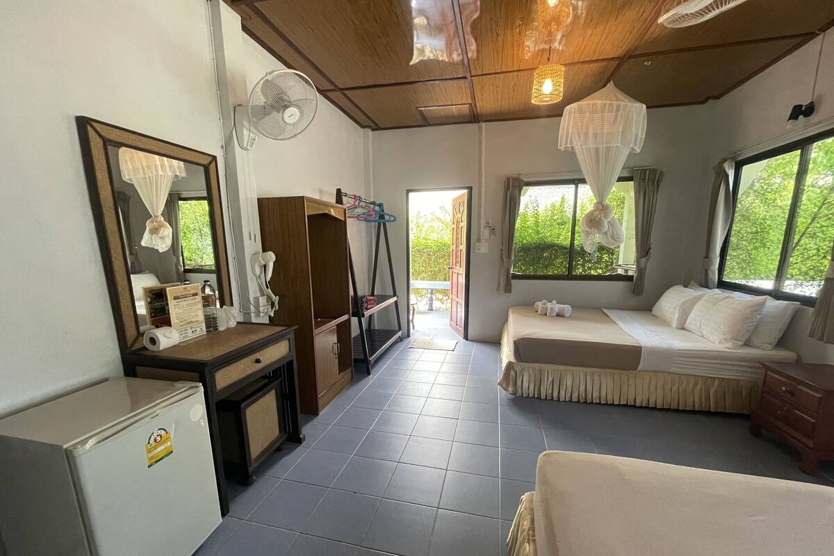 Una habitación con cama, escritorio, espejo, ventilador y vistas al exterior con vegetación.