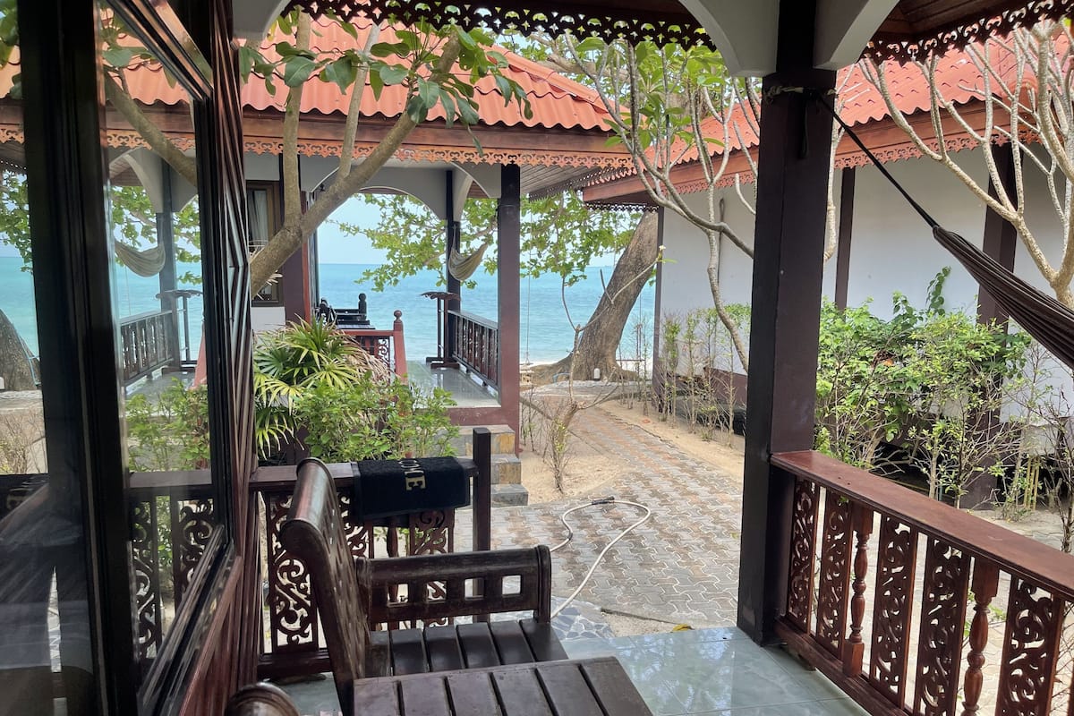 Una terraza de madera con un banco y una mesa, con vistas a una playa que cuenta con una estructura de techo rojo y vista al mar.