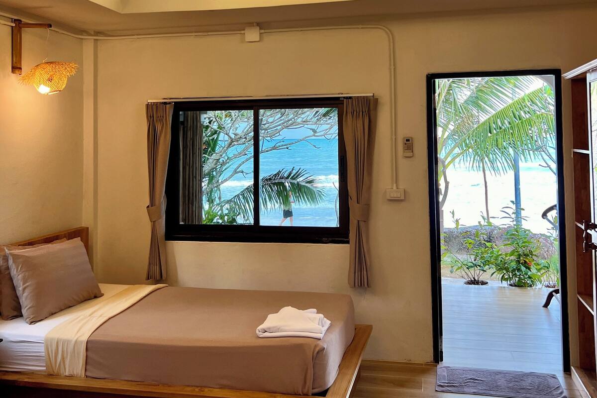 Un dormitorio con cama, mesita de noche, ventana con cortinas y vista a la playa.