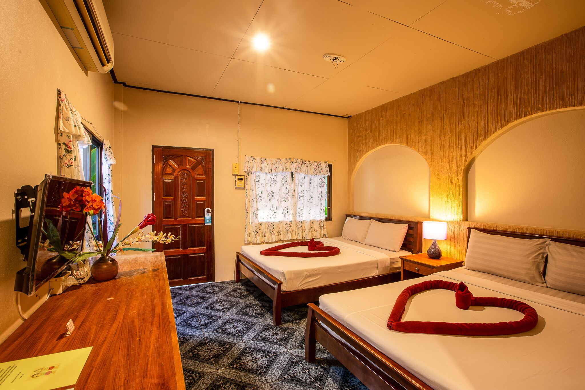 A hotel room with two beds, a wooden desk, a vase with flowers, and a heart-shaped towel on one bed.