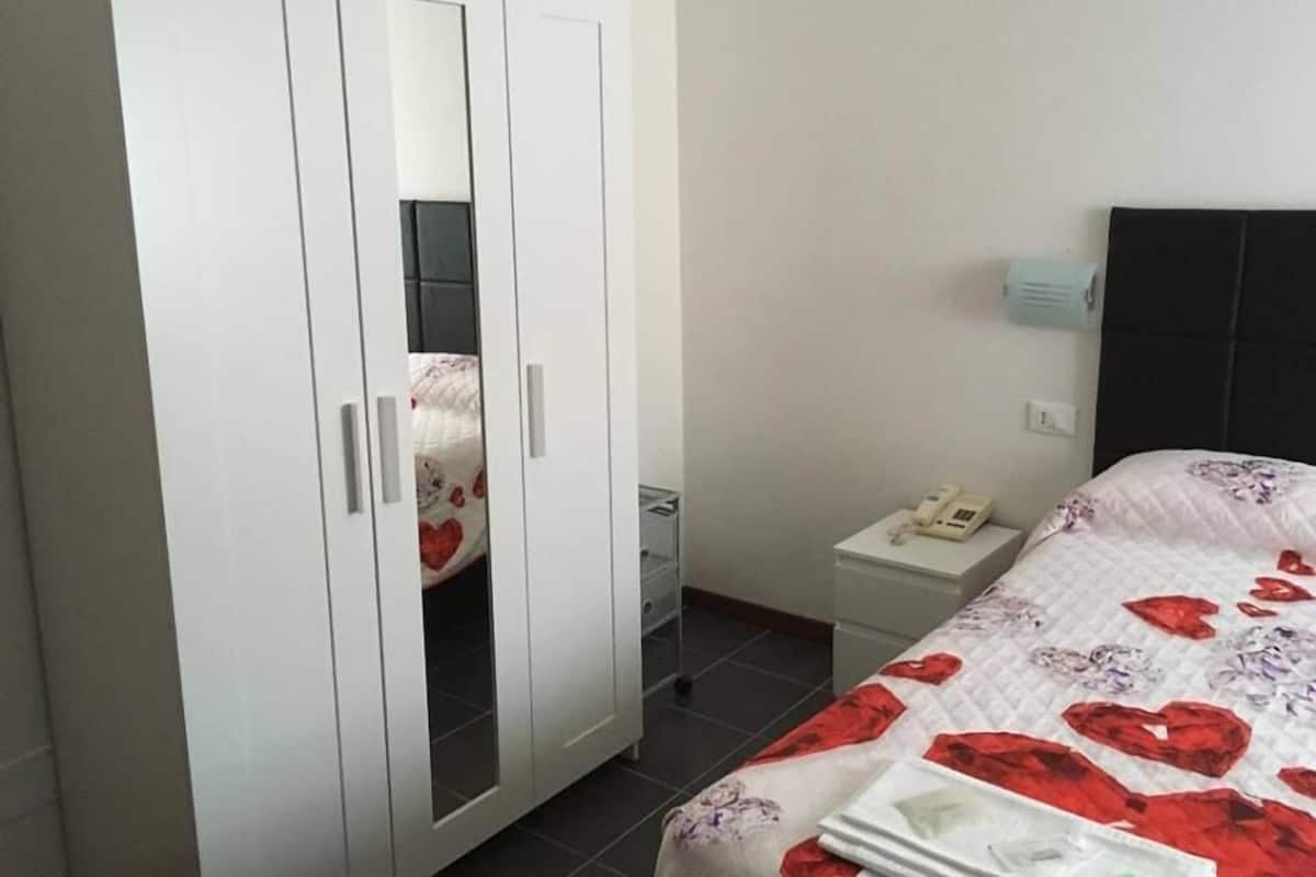 Un dormitorio con cama, un armario blanco, una mesita de noche con un teléfono y un calentador fijado a la pared.