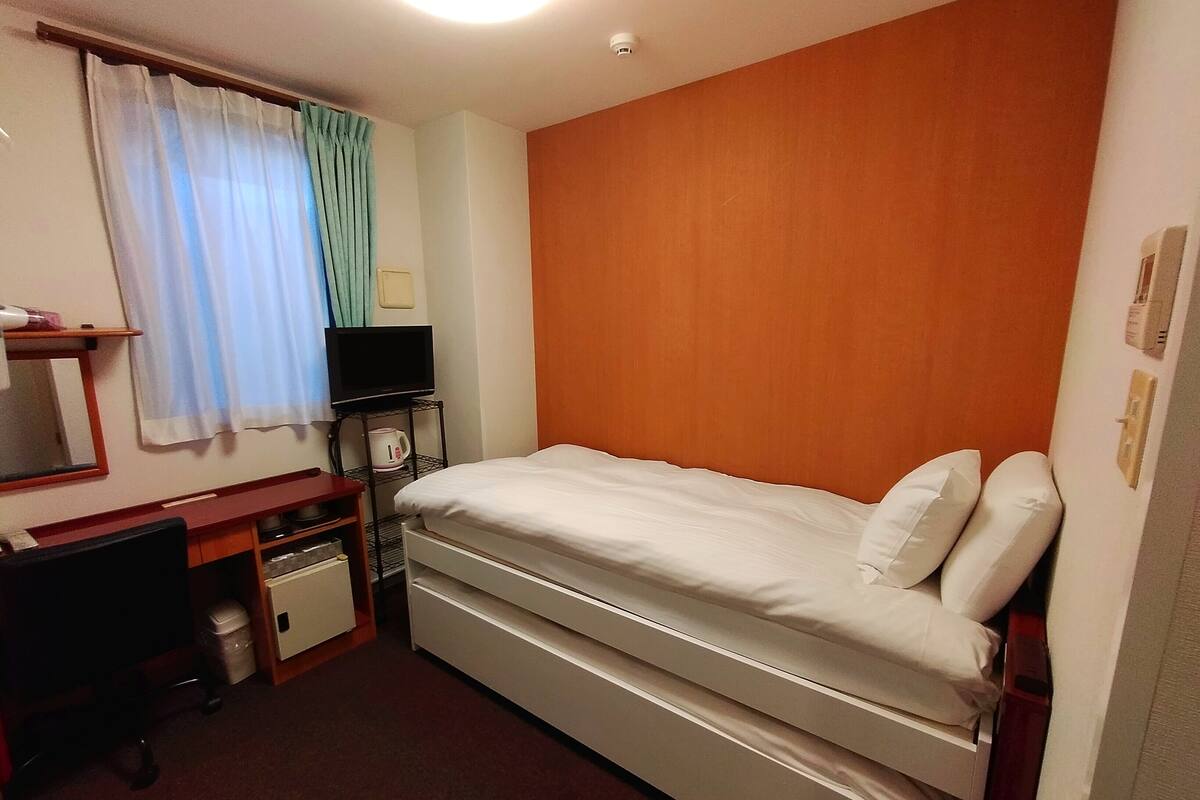Quarto de hotel com uma cama de solteiro, uma escrivaninha com cadeira, televisão e chaleira.