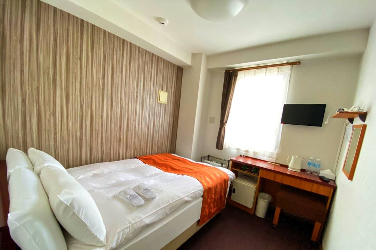 Quarto de hotel com cama, escrivaninha, cadeira, televisão e janela com cortinas.