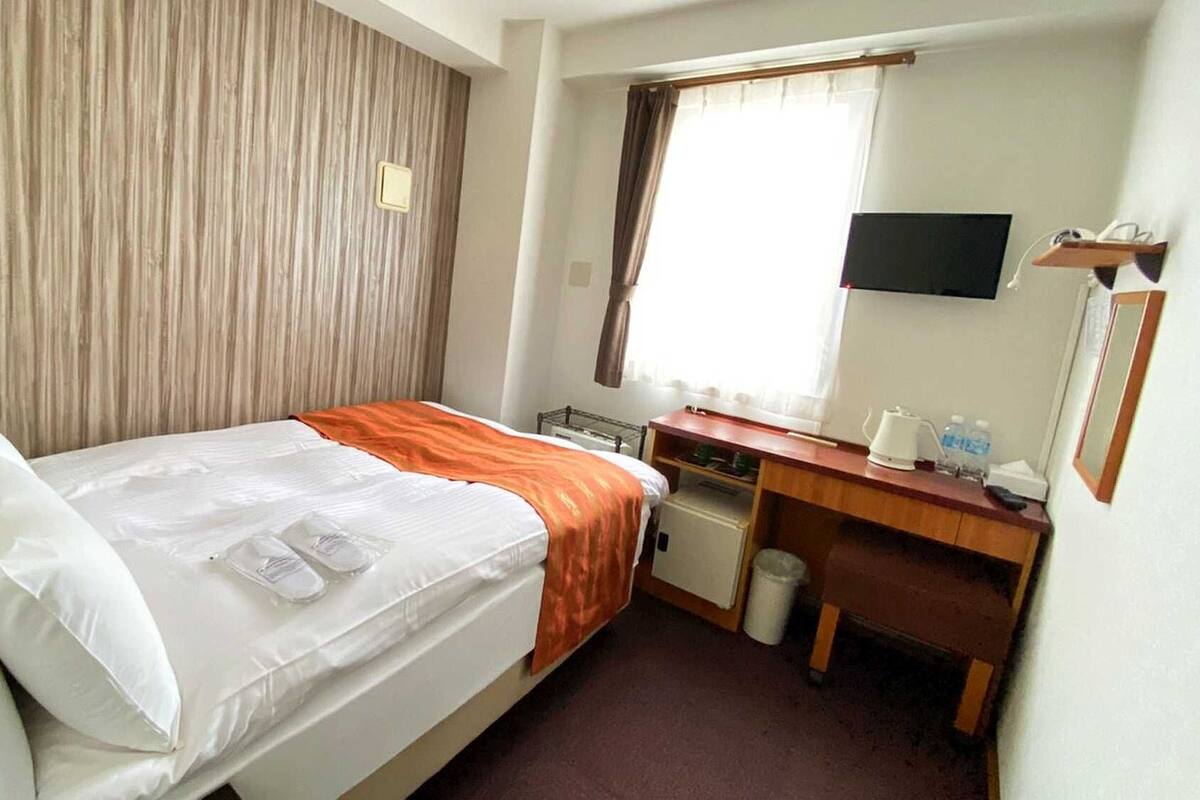 Quarto de hotel com cama, escrivaninha, cadeira, televisão e janela.