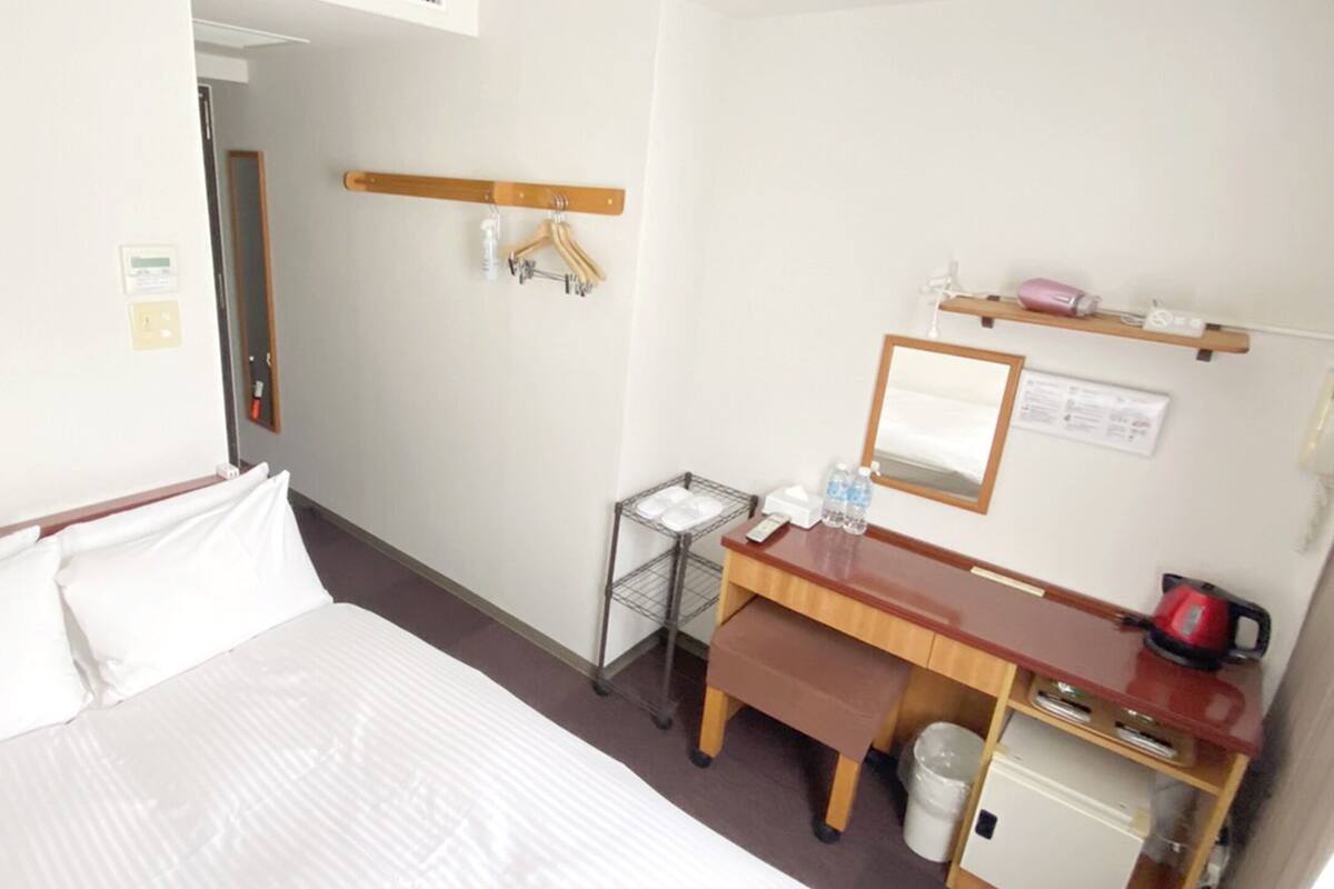 Quarto de hotel com cama, escrivaninha com espelho, lixeira pequena e prateleira com chaleira e garrafa de água.