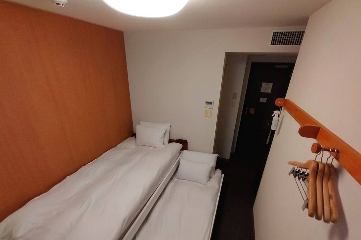 Quarto de hotel com uma cama de solteiro, cabeceira de madeira, porta-toalhas fixado na parede e uma porta que dá para outro quarto.