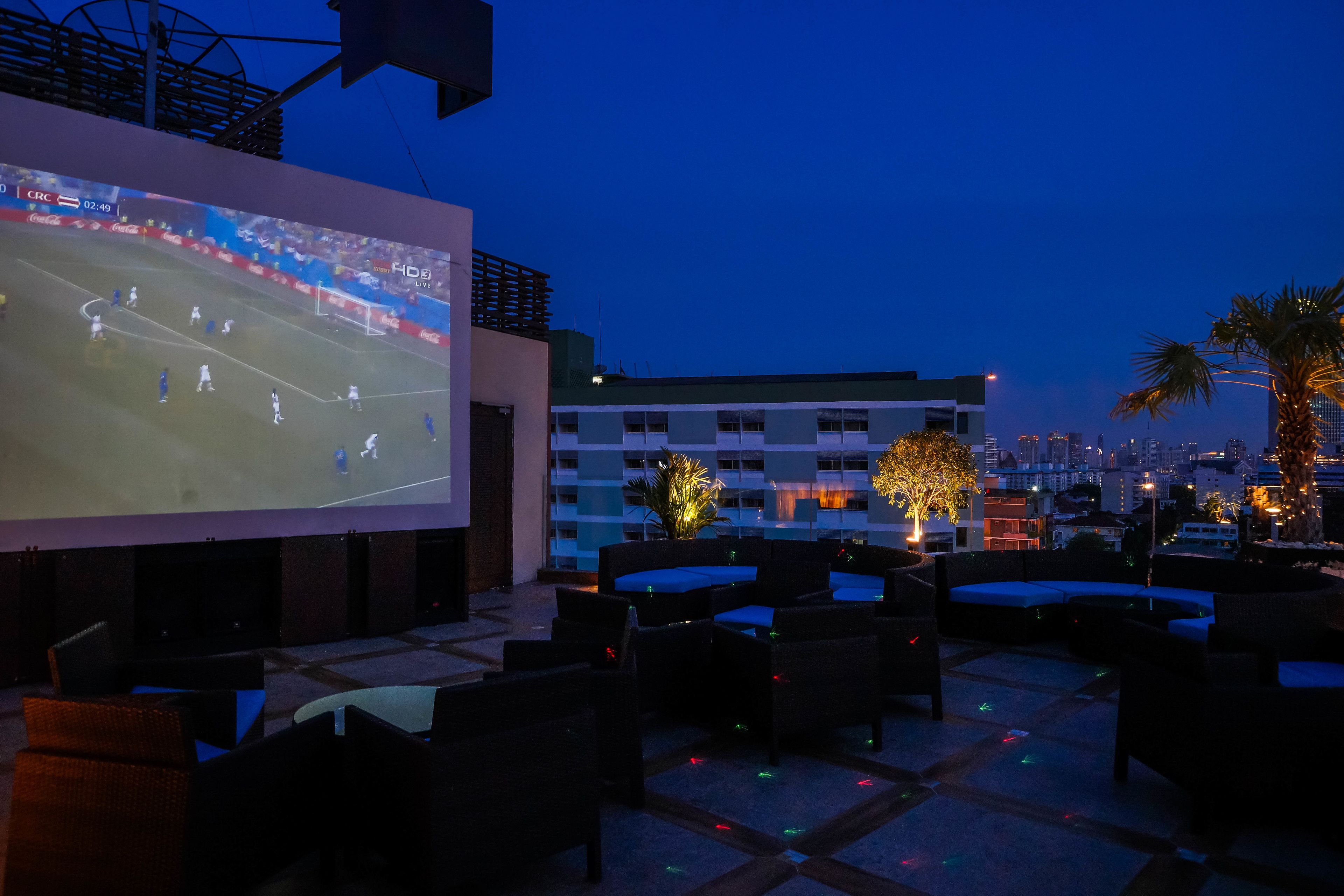 A rooftop bar with outdoor seating and a large screen displaying a soccer match.