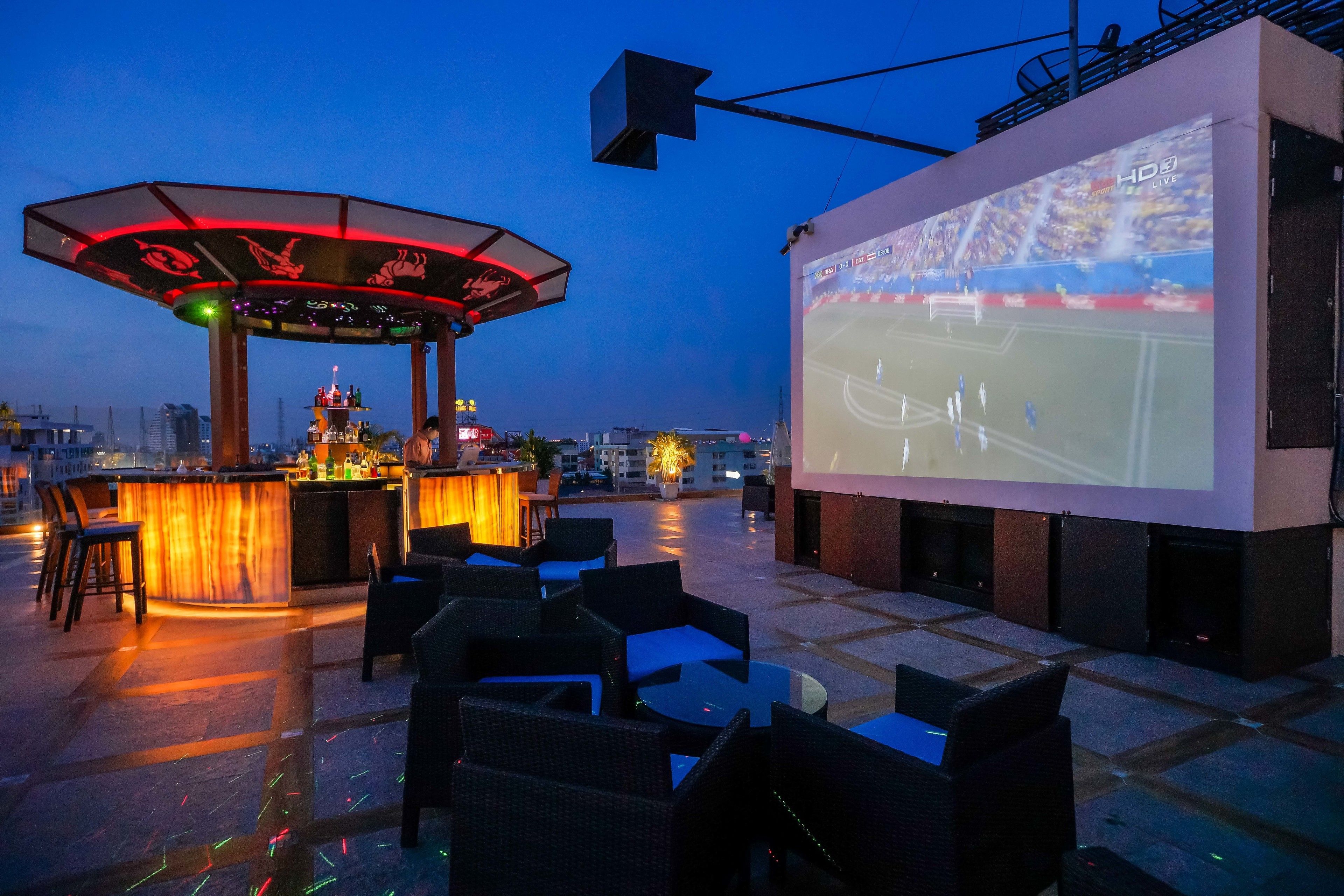 An outdoor bar area with a screen displaying a sports event, seating, and a bar counter.