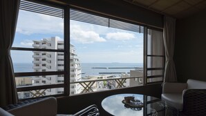 A room with a large window overlooking a cityscape and the sea. There is a round glass table with a decorative item on it, and two chairs. - Hotel Kanichi (Atami)