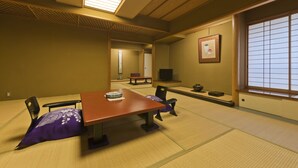 A traditional Japanese room with tatami flooring, a low table, chairs, a television, and a sliding door. - Hotel Kanichi (Atami)