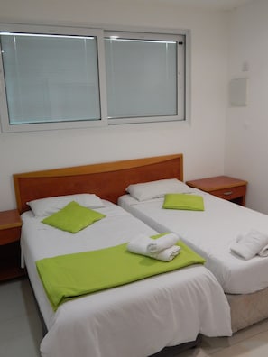 Habitación de hotel con dos camas individuales, cada una con una almohada verde y una toalla blanca. Se aprecia un cabecero de madera, una mesita de noche a cada lado de las camas y una ventana sobre ellas.