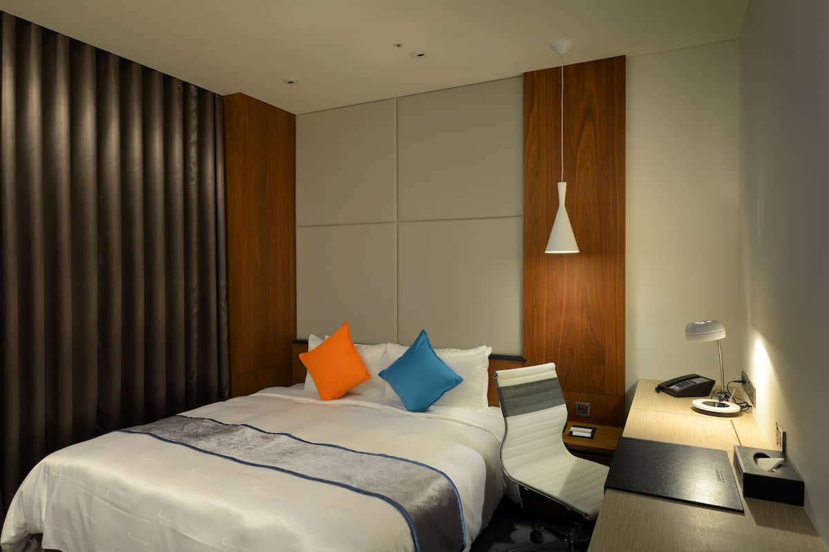 Una habitación de hotel moderna con una cama bien hecha, un escritorio con lámpara y un ventanal con cortinas.