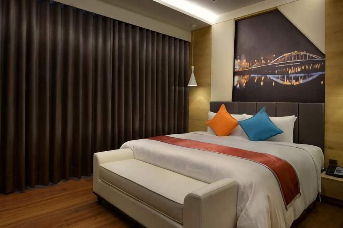 Habitación de hotel con cama, sofá, mesita de noche, lámpara y un cuadro de paisaje urbano en la pared.