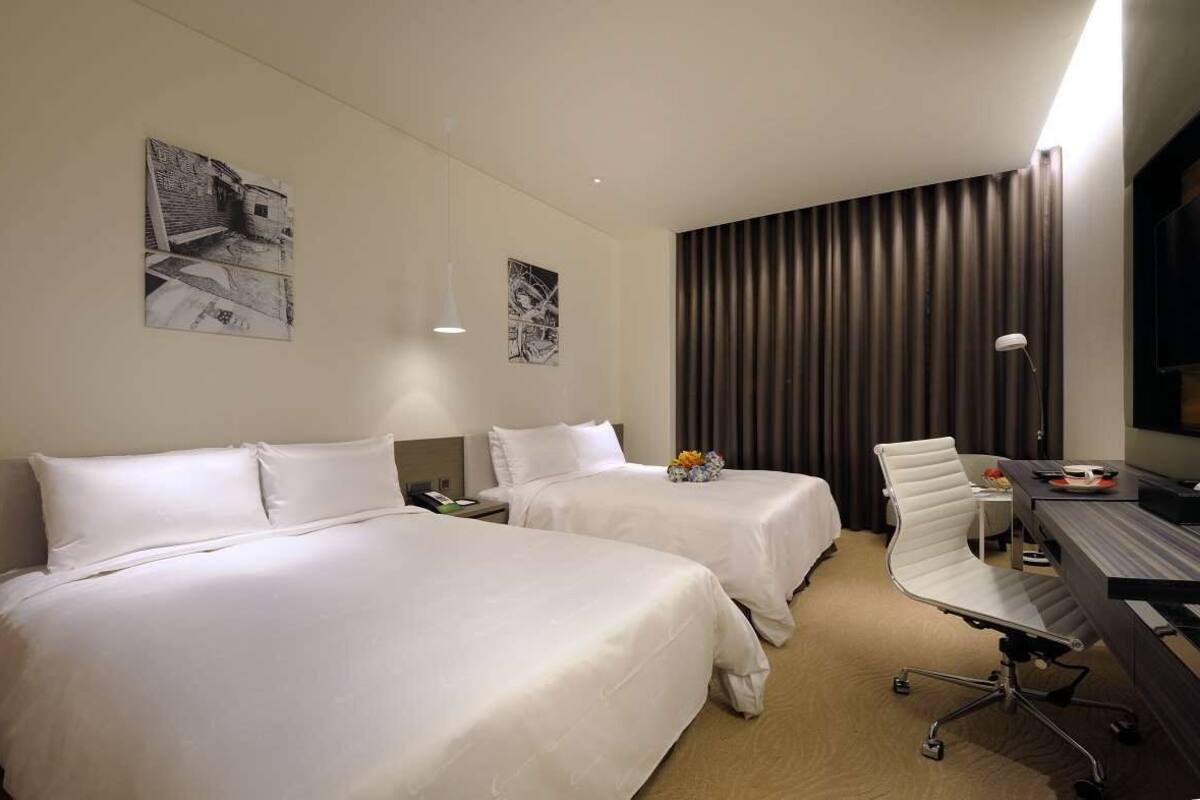 Habitación de hotel con dos camas, un escritorio, una silla, una lámpara y obras de arte en las paredes.