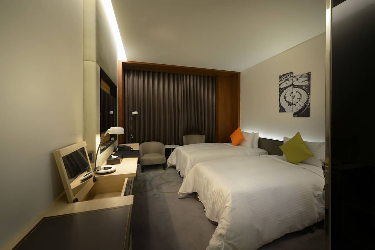 Habitación de hotel con dos camas, un escritorio con computadora, una silla, una mesita y un ventanal grande con cortinas.