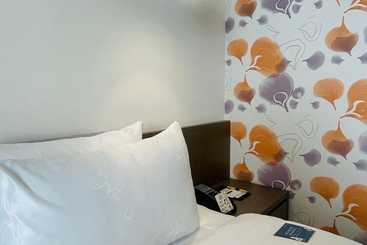 Habitación de hotel con cama, almohadas, mesita de noche, lámpara y pared con diseño de hojas.