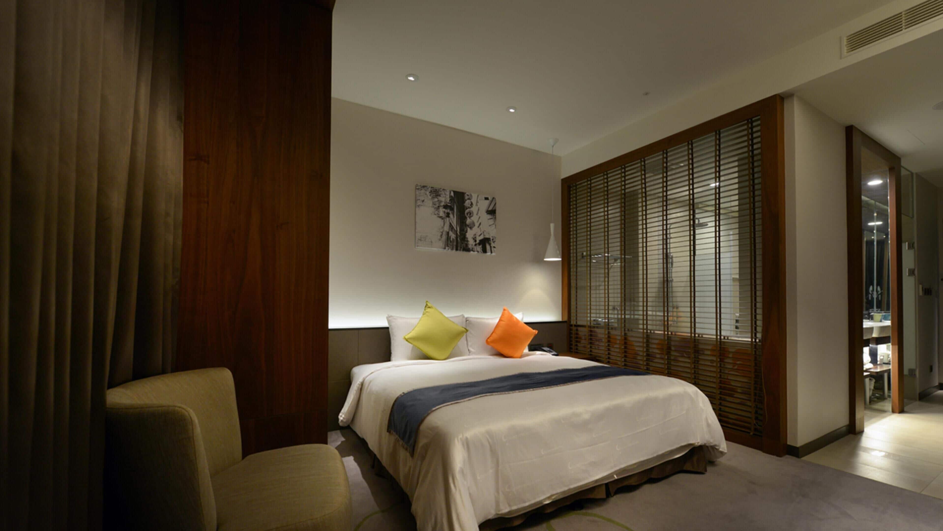 Ein Hotelzimmer mit einem Bett, einem Sessel, einem Nachttisch, einem Fenster mit Jalousien und einem Bild an der Wand.