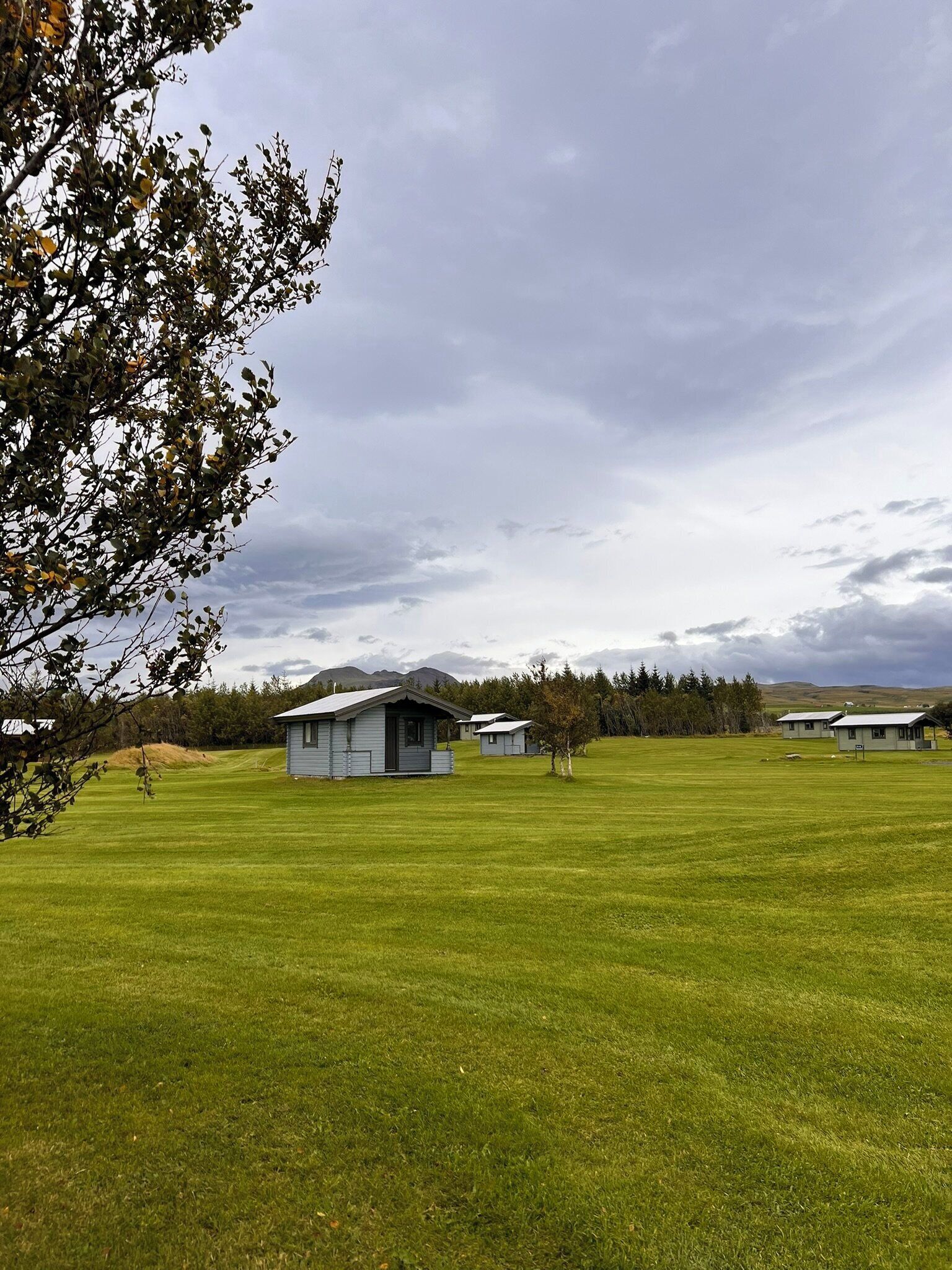 A grassy field with a small cabin and a larger building in the distance.