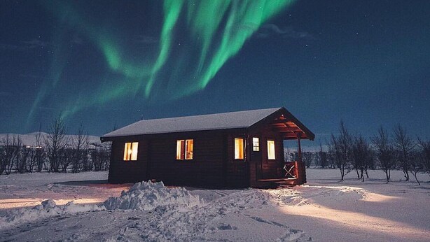 A cabin with a snow-covered ground and a view of the northern lights. - Hellisholar Cottages (Rangárþing eystra)