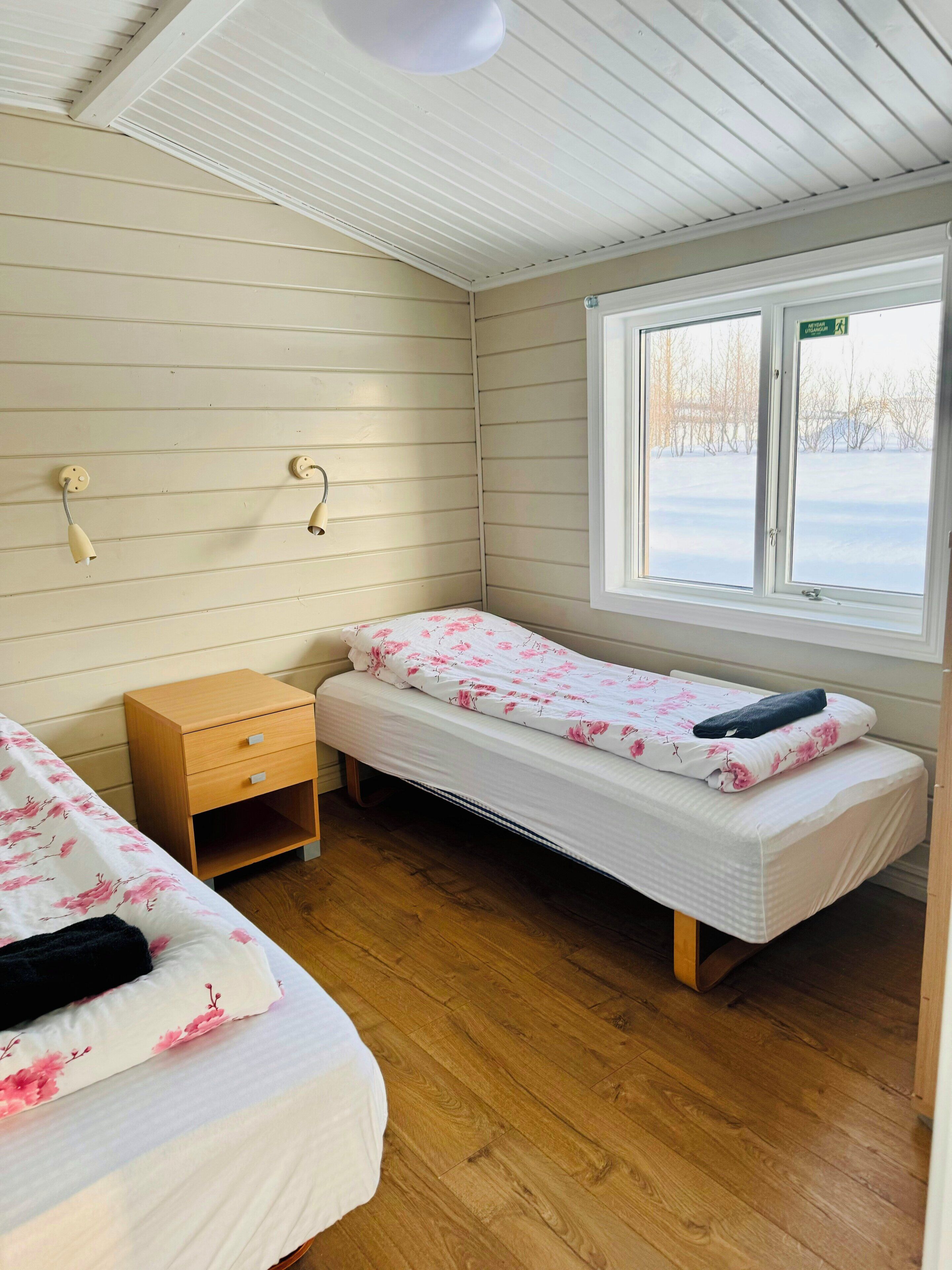 A small, single-room accommodation with two beds, a wooden nightstand, and a window with a view of a snowy landscape.