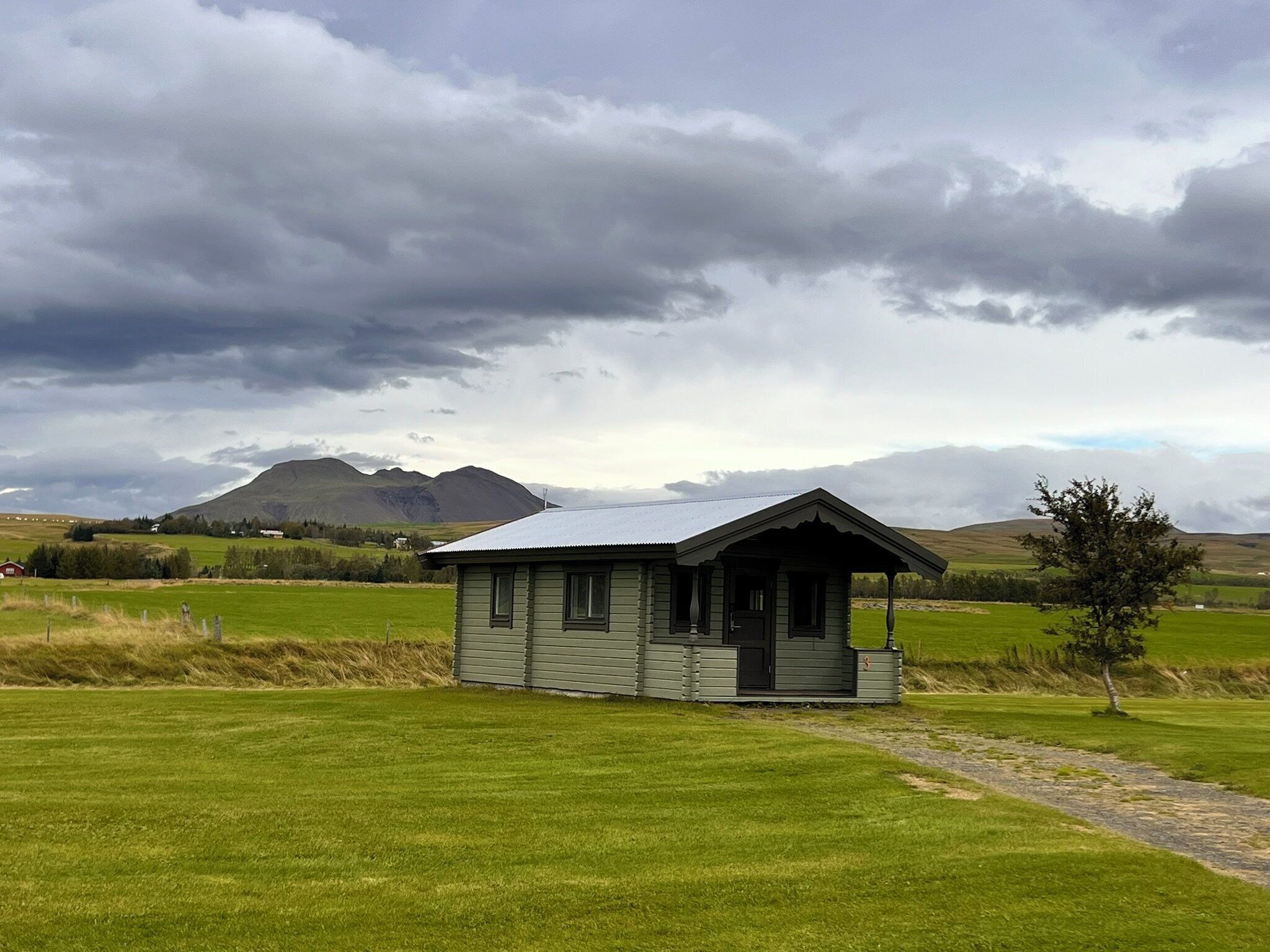 A small, single-story cabin with a porch, surrounded by a grassy field and mountains in the background.