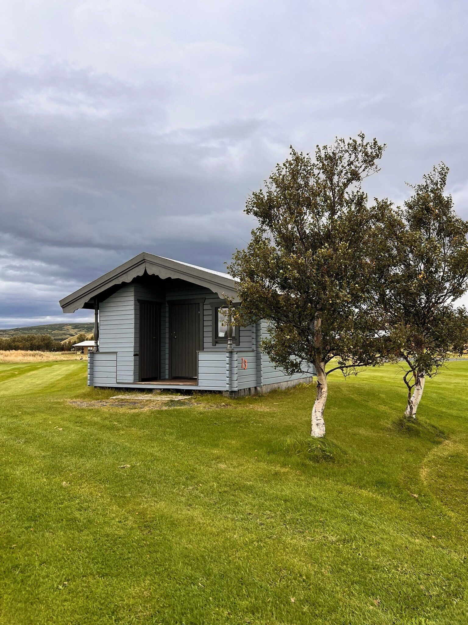 A small, single-story cabin with a dark roof and light siding, situated on a grassy field with a large tree nearby.