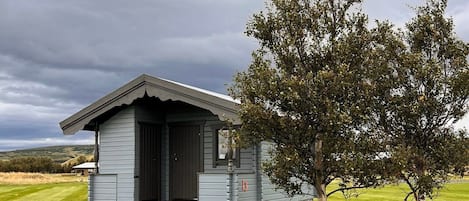 A small, single-story cabin with a dark roof and light siding, situated on a grassy field with a large tree nearby.