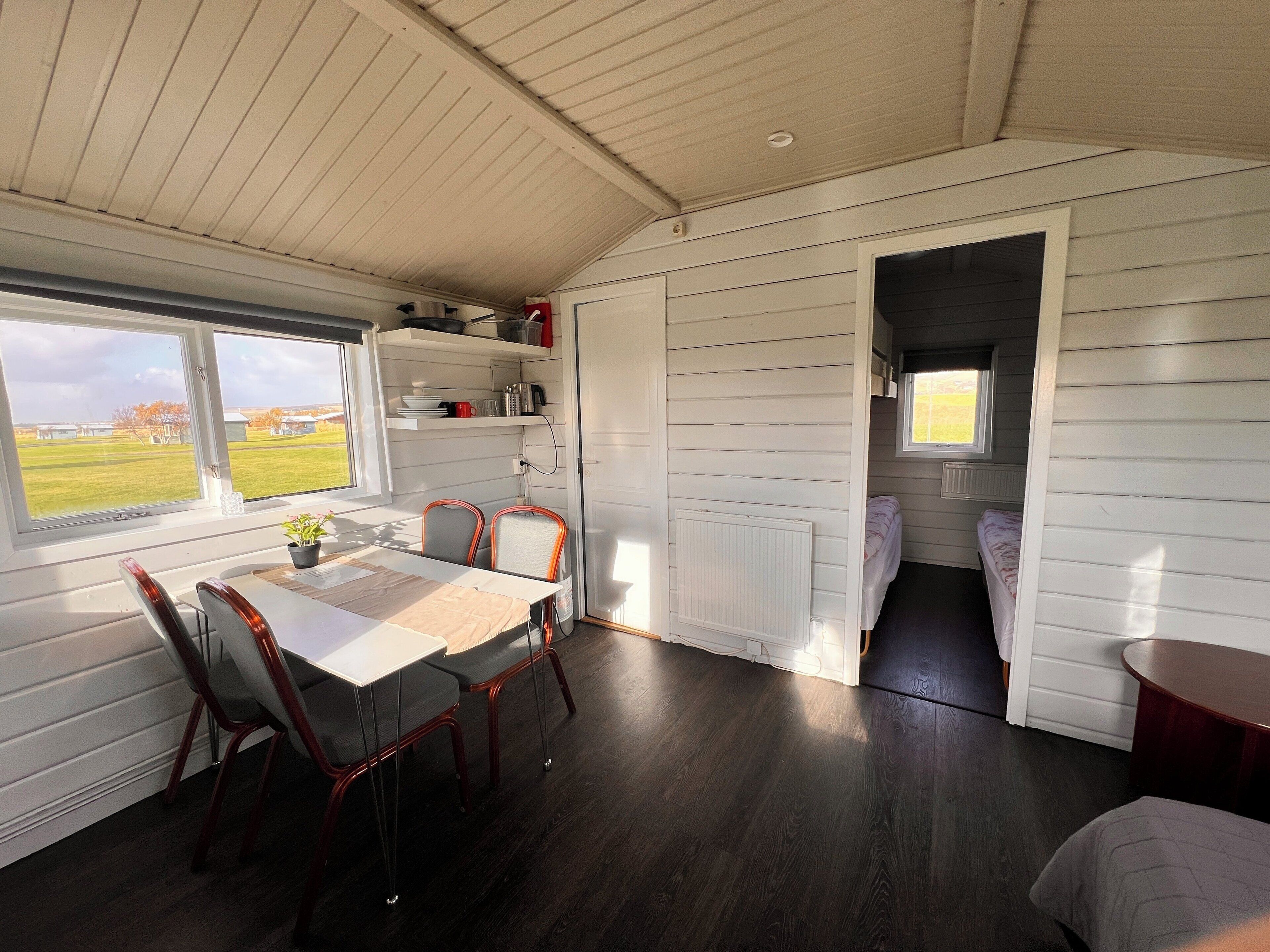 Habitación pequeña y luminosa, con mesa y sillas para comer, una kitchenette con estantes, y vistas al exterior.