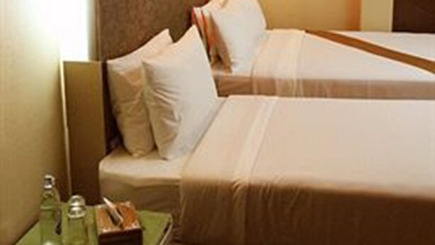 A hotel room with two beds, a nightstand with a bottle and tissue box, and a wall-mounted light. - Visiting Card Hotel & Resort (Bangkok)