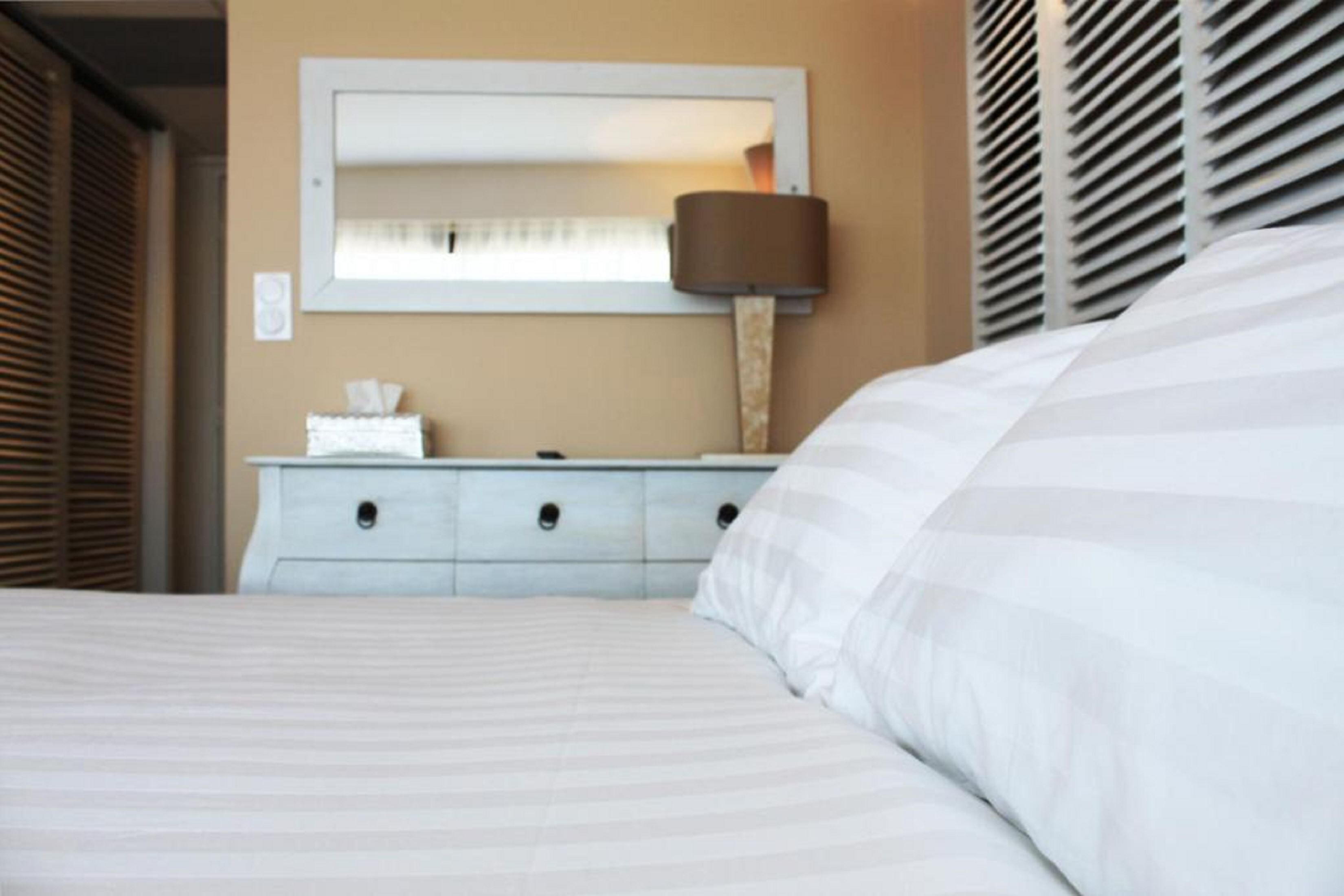 Een net opgemaakt bed met een gestreept dekbed, een nachtkastje met een lamp, en een spiegel aan de muur.