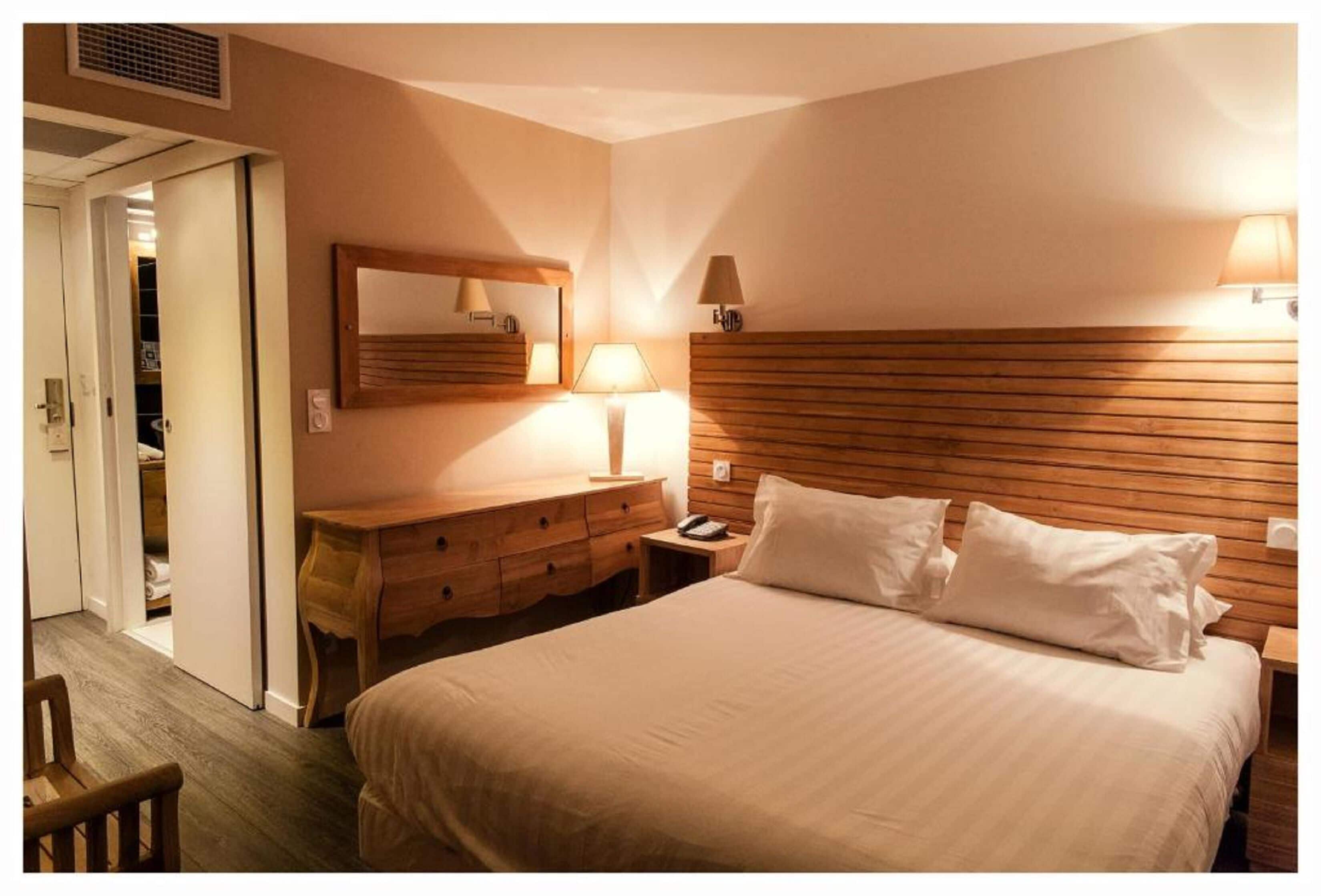 A hotel room with a large bed, wooden headboard, bedside lamps, a wooden nightstand, a mirror, and a wooden dresser.