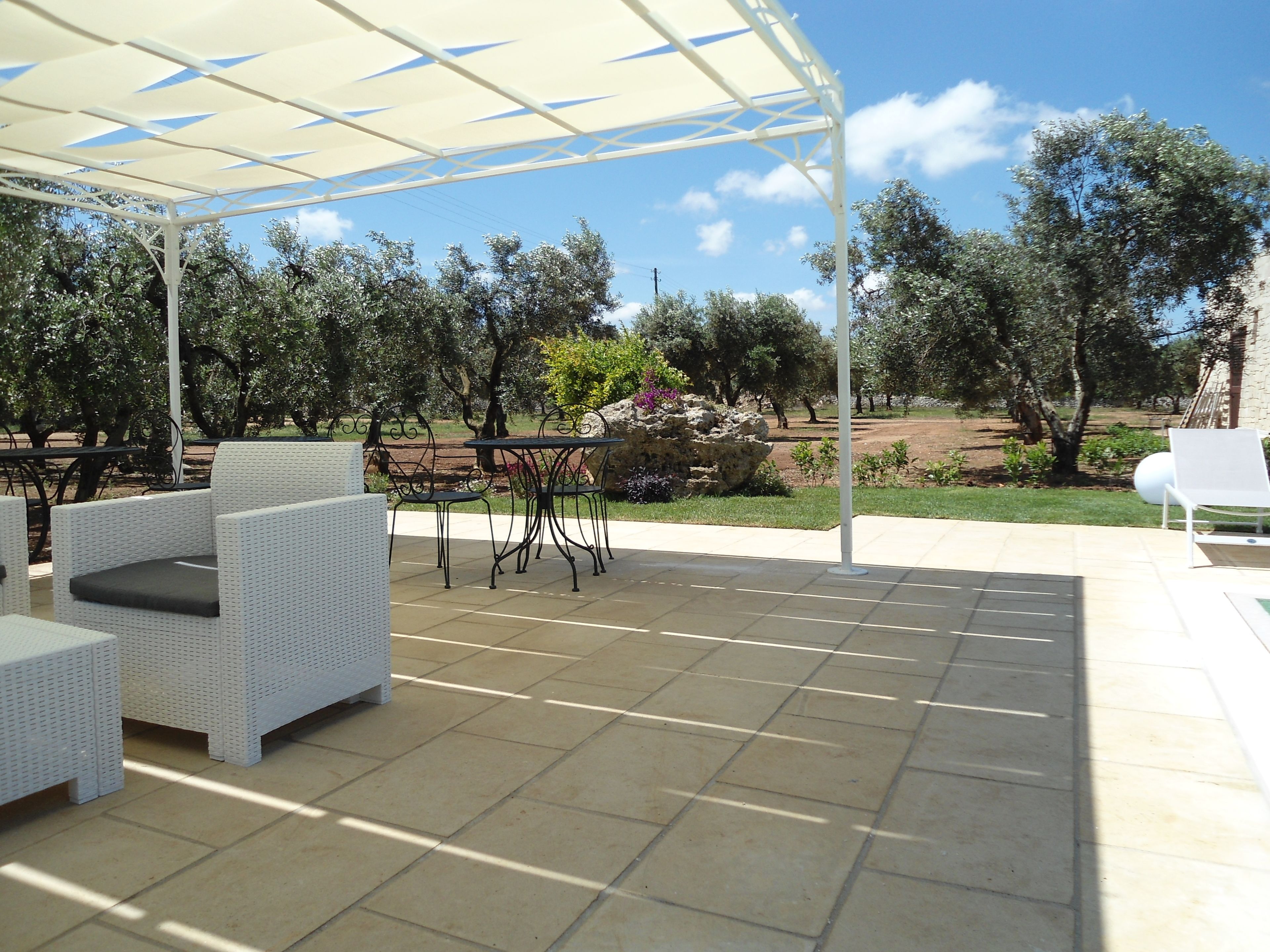An outdoor patio with white wicker furniture, a covered seating area, and a view of trees and a garden.