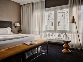 Ein Hotelzimmer mit einem großen Bett, einer Sitzbank, einer Lampe und Blick auf ein Gebäude durch das Fenster.