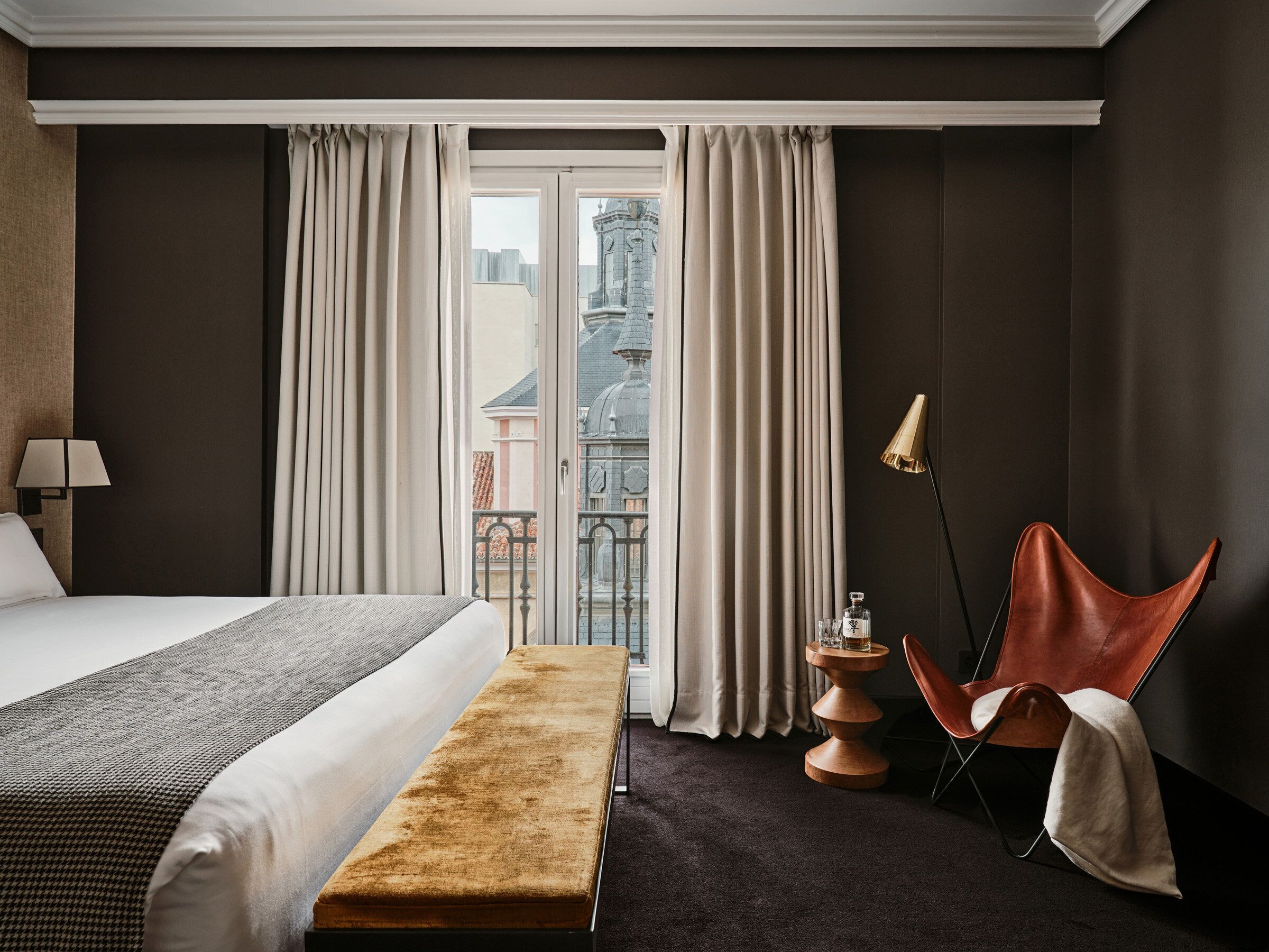 Habitación de hotel con cama, un banco, una silla, una lámpara y vistas de la ciudad a través de amplias ventanas.