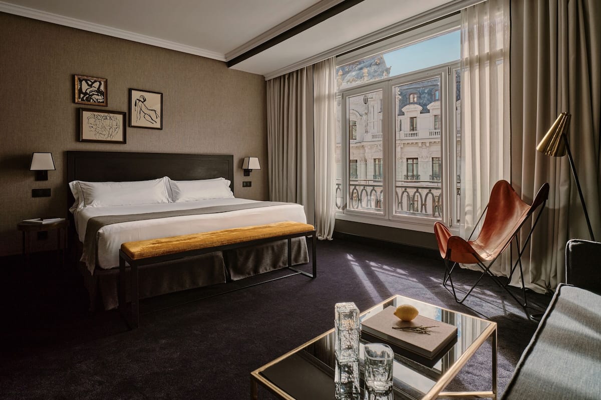 A hotel room with a large bed, a desk with a chair, a window with a view of buildings, and two wall-mounted lamps.