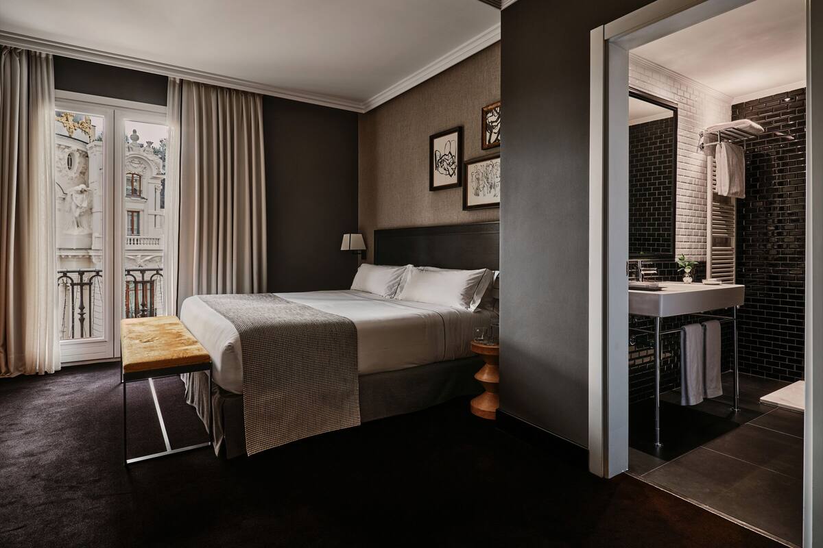 A modern hotel room with a large bed, a bedside table, a small round table, and a bathroom with a black tiled wall and a white sink.