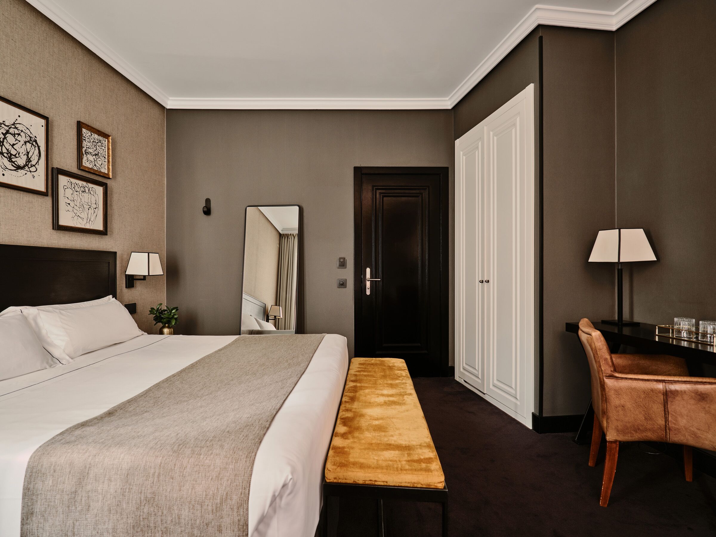 Une chambre d’hôtel avec un grand lit, un banc, un bureau avec une chaise, un miroir et des œuvres d’art encadrées accrochées au mur.