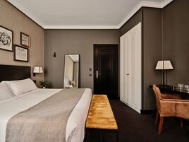 Una camera d'albergo con un grande letto, una panca, una scrivania con una sedia, uno specchio e quadri appesi al muro.