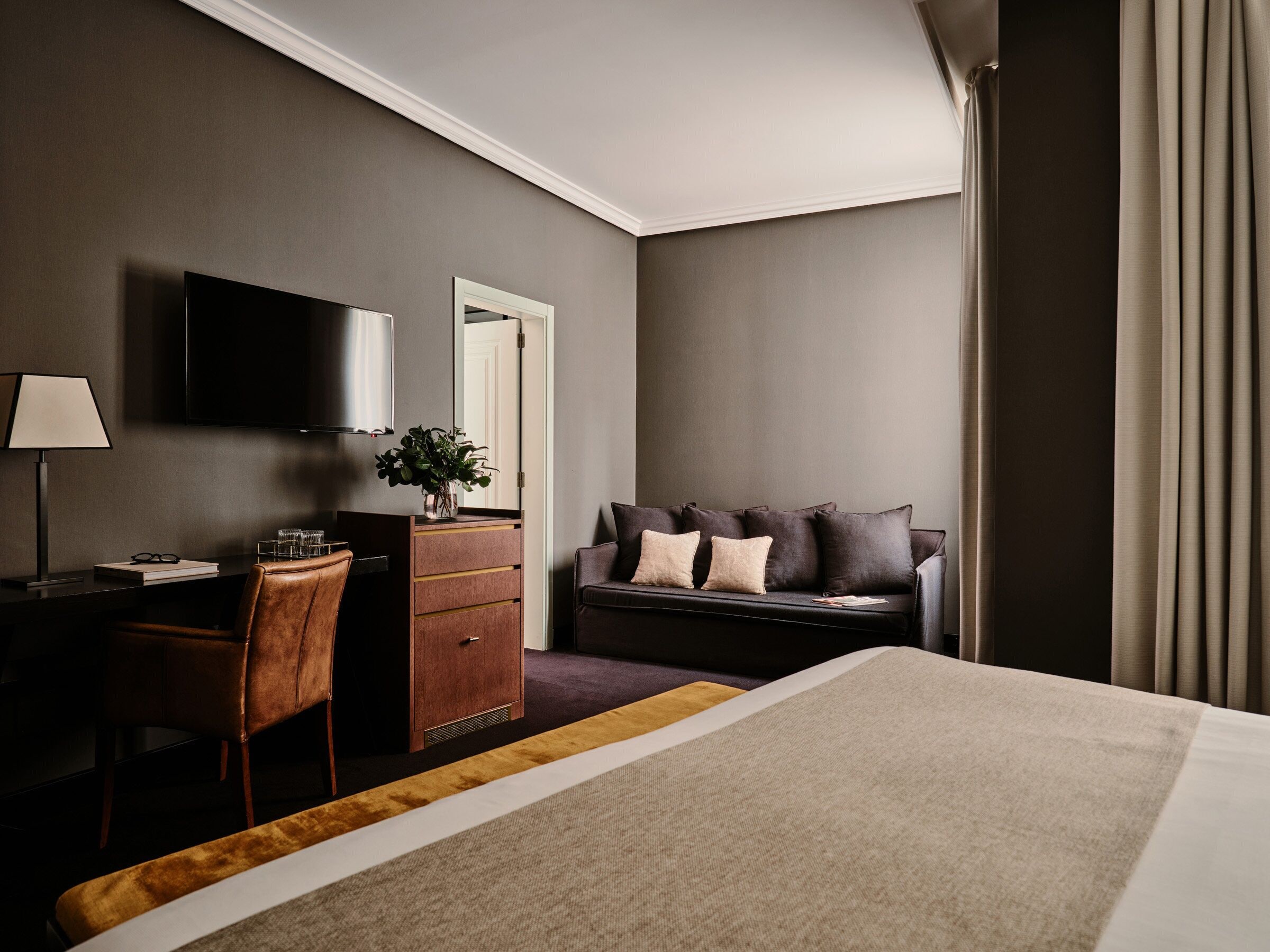 A hotel room with a bed, a desk with a lamp, a chair, a sofa, and a TV.