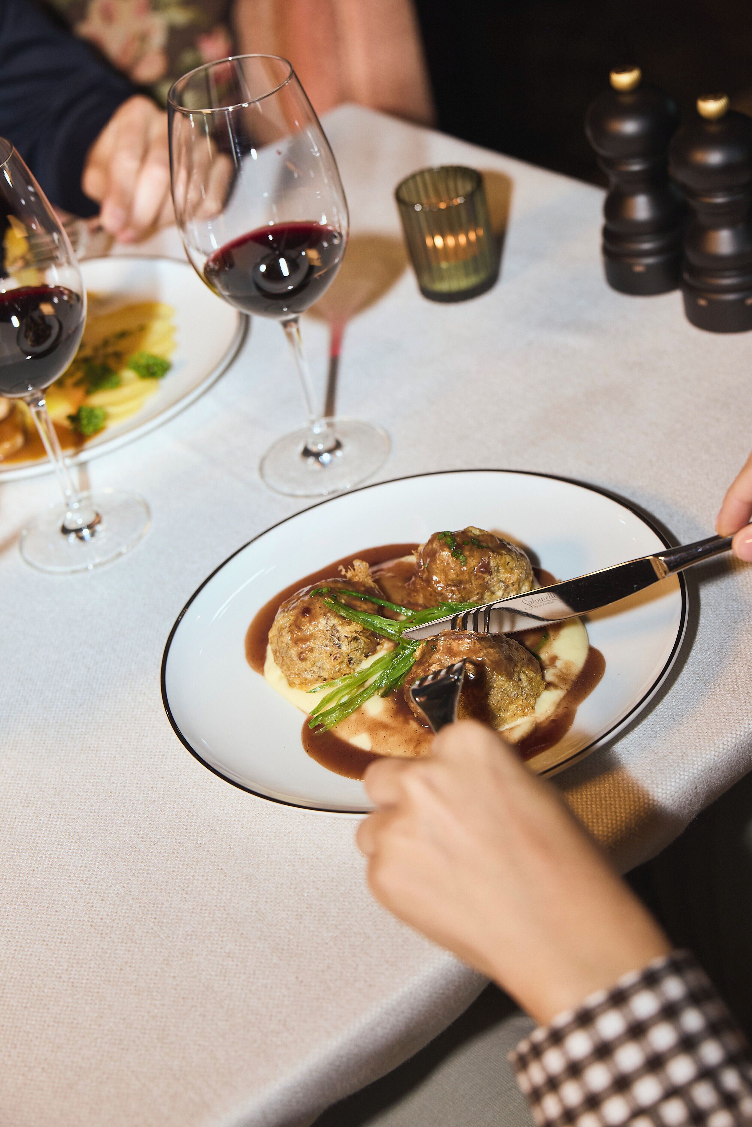 A dining table with a plate of food, a glass of red wine, and a person using a knife and fork.